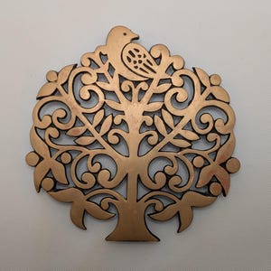 Peut inclure: Un objet décoratif en forme d'arbre de couleur bronze avec des motifs découpés complexes. Un oiseau est perché au sommet de l'arbre. L'arbre présente des branches tourbillonnantes, des feuilles et un tronc détaillé. L'objet est probablement en métal et a un aspect rustique.
