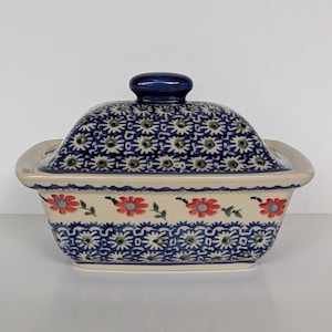 Op de afbeelding: Een rechthoekige keramische botervloot met deksel, voorzien van een crèmekleurige basis versierd met een patroon van kleine blauwe en witte bloemmotieven en een horizontale band van rode bloemen met groene bladeren rond het midden.