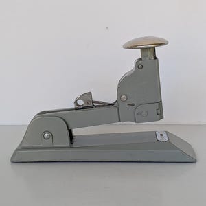 May include: A vintage gray stapler with a silver top button. The stapler is made of metal and has a classic design. It is sitting on a white surface.