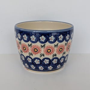 May include: A ceramic bowl with a cream-colored interior and a blue exterior decorated with a repeating floral pattern. The pattern features red and yellow flowers with green leaves, and white flower-shaped accents.