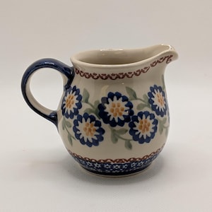 May include: Cream-colored ceramic creamer with a dark blue handle and spout. The creamer is decorated with blue and yellow floral patterns and green leaves. A dark blue and brown border accents the top and bottom.