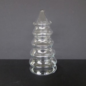 Puede incluir: Un objeto decorativo transparente en forma de árbol de Navidad. El árbol está compuesto por cinco secciones redondeadas apiladas, con una parte superior puntiaguda. El vidrio es transparente, permitiendo el paso de la luz. El árbol se encuentra sobre una superficie oscura.