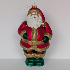May include: A Christmas ornament shaped like Santa Claus. The ornament features a red coat with gold trim, a white beard, and green boots. A gold loop is attached to the top for hanging. The ornament is a festive decoration for the holiday season.