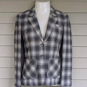 May include: A gray, black, and white plaid blazer with two front pockets and a single-button closure. The blazer is displayed on a white mannequin against a light-colored wooden background. The jacket has a classic, tailored design.