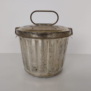 Op de afbeelding: Een zilveren metalen cakevorm met een geribbeld ontwerp en een scharnierend deksel met een draadhandvat. De vorm heeft een versleten, vintage uitstraling.