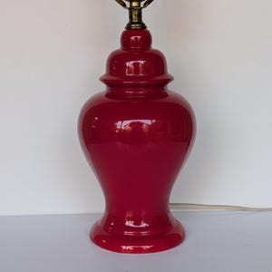 May include: A glossy, deep red table lamp with a rounded, bulbous body and a narrow neck. The lamp features a metallic gold-toned socket assembly at the top, designed to hold a light bulb, and includes a thin white electrical cord extending outward.