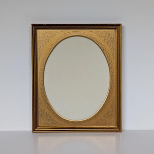 May include: A rectangular gold picture frame with a dark brown outer edge and an oval inner opening. The frame features an embossed gold matting with intricate floral scrollwork patterns in each corner and along the oval border.
