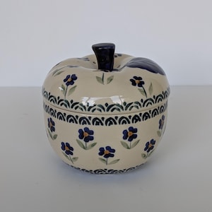 Könnte beinhalten: Ein keramikapfelförmiges Gefäß mit Deckel. Das Gefäß ist cremefarben mit einem Muster aus blauen Blumen und grünen Blättern. Der Deckel hat einen dunkelblauen Stiel. Das Gefäß wird wahrscheinlich zur Aufbewahrung oder Dekoration verwendet.