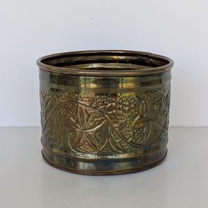 May include: A cylindrical brass planter with an embossed floral design. The planter has a dark patina and a slightly flared rim. The detailed pattern includes leaves and vines, adding a vintage aesthetic. Suitable for indoor or outdoor use.