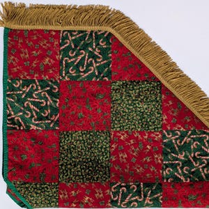 May include: A Christmas tree skirt with a patchwork design. The skirt features red and green squares with festive patterns like candy canes and poinsettias. It has a gold fringe trim.