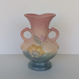 May include: A vintage ceramic vase with a two-tone color scheme. The top is a soft pink, transitioning to a cream and blue base. It features a floral design with yellow and purple accents, and two loop handles.