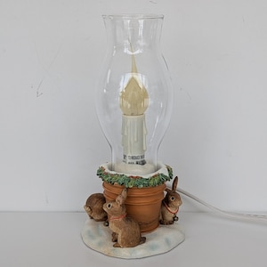 May include: A decorative lamp featuring a clear glass shade and a candle-shaped light bulb. The base is a terracotta pot with three brown bunny figurines and a garland of green leaves. The lamp is on a white surface.
