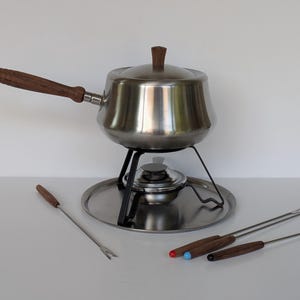 Op de afbeelding: Een roestvrijstalen fonduepan met houten handvat en dekselknop, rustend op een zwarte metalen standaard met brander. De set bevat een ronde roestvrijstalen onderplaat en vier metalen fonduevorken met houten handvatten en gekleurde uiteinden.