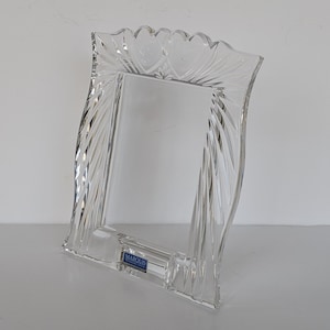 May include: A clear, decorative glass picture frame with a scalloped top edge. The frame has a rectangular opening and is embellished with vertical and curved etched lines. A small blue and white label is visible on the bottom.