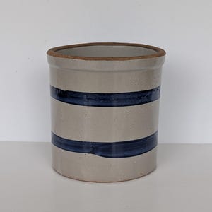 May include: A cylindrical stoneware crock with a light gray body and two dark blue horizontal stripes. The rim is a darker brown color. This vintage kitchenware item is suitable for storage or display.