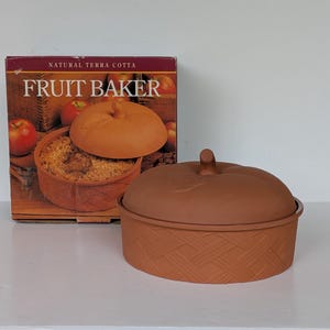 May include: A round, natural terra cotta fruit baker with a matching lid featuring a stem-like handle. The exterior of the base has a textured, woven basket-weave pattern. The product is displayed in front of its original box labeled FRUIT BAKER.