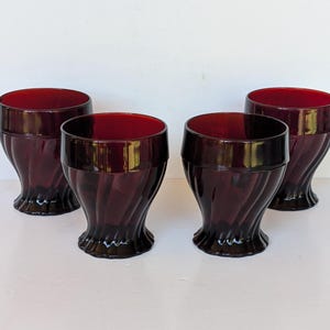 Könnte beinhalten: Ein Set aus vier dunkelroten Glasbechern mit einem glatten oberen Rand und einem unteren Bereich mit vertikalen, geschwungenen Rillenmustern, die bis zu einem ausgestellten, gewellten Boden verlaufen und ein einheitliches Design bilden.