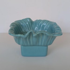 May include: A light blue ceramic bowl with a square base and a wide, ruffled, fluted rim. The bowl features a glossy, solid-colored finish throughout, with vertical ridges extending from the center toward the wavy, scalloped edges of the opening.