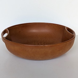 Pode incluir: Uma tigela oval marrom com duas alças. A tigela tem um interior liso e um exterior texturizado. As alças são integradas no design da tigela. Adequada para servir ou expor. Medidas em centímetros.