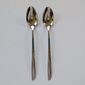 May include: Two stainless steel spoons with oval bowls and long, slender handles. The spoons reflect the surrounding environment. The handles have a decorative starburst design near the base. The spoons are set against a white background.
