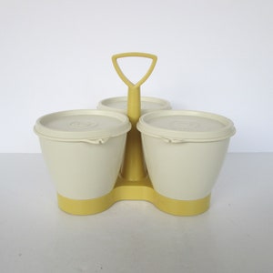 以下が含まれることがあります： クリーム色と黄色のレトロな調味料入れ。蓋付きの3つの個別の容器があり、黄色のハンドルでまとめられています。調味料入れはプラスチック製で、スパイスやその他の小物を入れるように設計されています。高さは約15cmです。