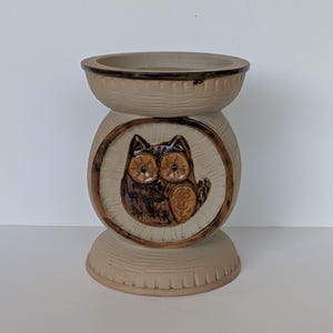 May include: A beige ceramic owl planter with a brown owl design in a circular frame. The planter has a textured surface and a wide top opening. The owl has large eyes and detailed feathers.