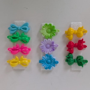 May include: A collection of colorful hair clips, including bows and flower designs. The clips are in various bright colors such as blue, green, pink, yellow, red, and purple. They are arranged on white foam.