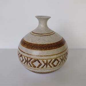 May include: A ceramic vase with a round body and a narrow neck. The vase is primarily beige with brown horizontal stripes and a geometric pattern near the base. The vase has a matte finish and a simple, elegant design.