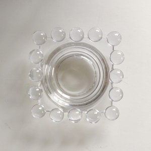 May include: Clear glass ashtray with a square frame of clear, round glass beads. The ashtray has a circular indentation in the center. The design is simple and modern, suitable for a minimalist aesthetic.