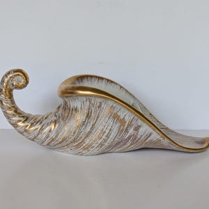May include: A cornucopia-shaped ceramic vessel featuring a white base with a textured gold-tone finish. The piece has a curved, tapered tail and a wide, open mouth with a smooth gold-painted rim, showcasing a distinct brushed metallic pattern.