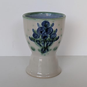 Puede incluir: Un jarrón de cerámica con base acampanada y boca ancha. El jarrón es de color blanquecino con un diseño floral azul y verde. El borde es de un color azul-verdoso más oscuro. El jarrón es probablemente hecho a mano y tiene una estética rústica.