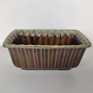 May include: A brown and green ceramic planter with a square shape and a fluted design. The planter has a light green glaze on the top edge and a darker brown glaze on the bottom.