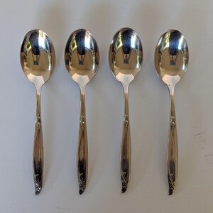May include: A set of four matching silver-toned metal spoons arranged in a vertical row. Each spoon features a polished, reflective oval bowl and a slender handle decorated with a subtle, embossed floral scroll pattern at the base of the stem.