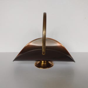 May include: A copper and brass log holder with a curved handle. The holder has a round base and a curved, half-circle shape.