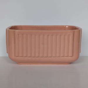 May include: A rectangular ceramic planter with a glossy, muted peach finish. The exterior features a series of vertical, raised ridges along the front and sides, creating a textured, linear pattern. The planter has rounded corners and a flat base.