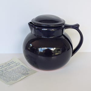 Può includere: Una caraffa da caffè in ceramica blu scuro con coperchio abbinato e manico curvo. La caraffa ha un corpo arrotondato e una finitura lucida. Un piccolo foglio di carta con testo si trova nell'angolo in basso a sinistra. La caraffa è progettata per 12 tazze.