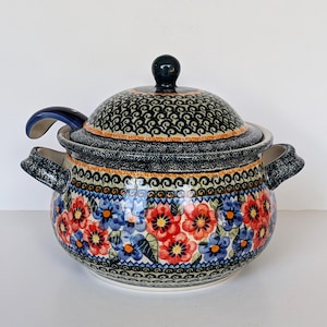 May include: A ceramic soup tureen with a matching lid and side handles, featuring a vibrant pattern of red and blue flowers with green leaves on a cream background, accented by dark blue and black decorative borders and a dark blue ladle handle.