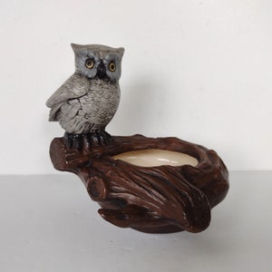 May include: A ceramic owl planter with a brown tree branch base. The owl is grey with yellow eyes and is perched on the branch. The planter has a small, round opening for planting.