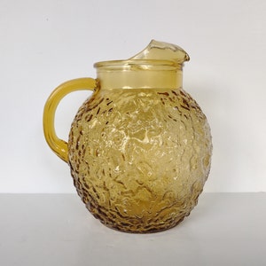 May include: A vintage amber glass pitcher with a textured surface. The pitcher has a round body and a handle.