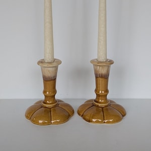 Puede incluir: Dos candelabros de cerámica con velas beige. Los candelabros son de color marrón cálido con una base en forma de flor. Los candelabros tienen un diseño degradado beige y marrón. Las velas son de color beige claro.