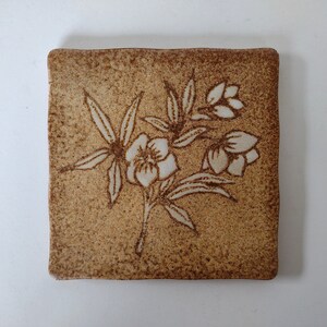 May include: Square ceramic tile with a textured, speckled brown surface. A floral design in off-white is etched into the tile, featuring stylized flowers and leaves. The tile has slightly rounded edges and a rustic aesthetic.