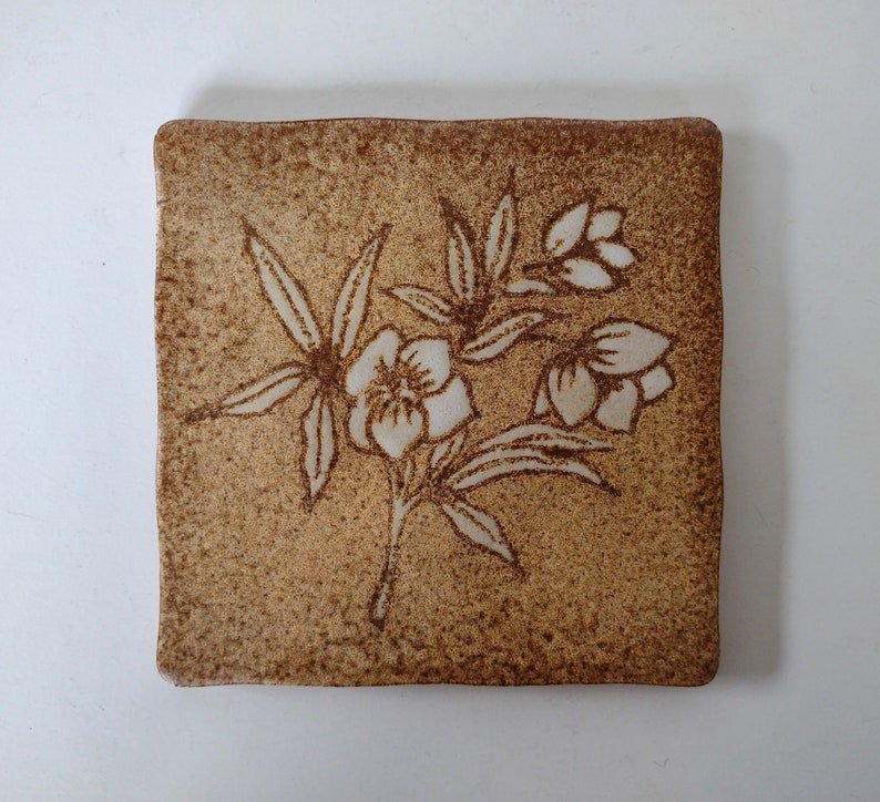 May include: Square ceramic tile with a textured, speckled brown surface. A floral design in off-white is etched into the tile, featuring stylized flowers and leaves. The tile has slightly rounded edges and a rustic aesthetic.