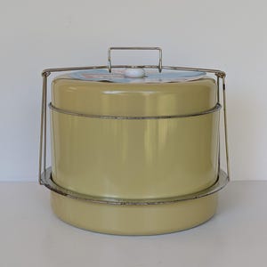 May include: A cylindrical, pale yellow metal cake carrier with a smooth finish. It features a silver-toned metal locking handle mechanism that extends over the top, secured by a white knob. A circular paper label is visible on the lid.