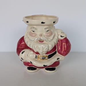 May include: A ceramic Santa Claus figurine with a cream-colored face, beard, and hat. The Santa figure has rosy cheeks and a red suit with black and gold accents. He holds a red sack and has a circular opening at the top.