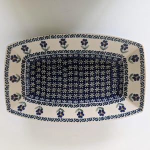 May include: A rectangular ceramic serving dish featuring a cream-colored rim decorated with a repeating pattern of blue flowers and green leaves. The deep center of the dish is filled with a dense, intricate pattern of small white star-shaped flowers.