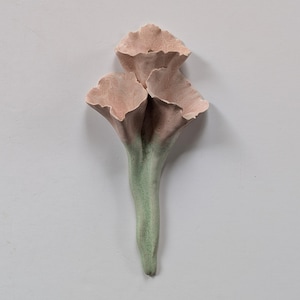 May include: A wall-mounted ceramic sculpture featuring three trumpet-shaped flowers with ruffled edges. The petals are a soft, dusty pink color, while the elongated stems transition into a muted, pale sage green at the base, creating a floral form.