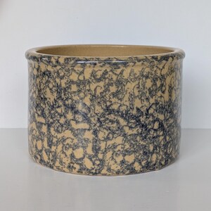 May include: A cylindrical ceramic crock featuring a tan base color with a dark blue sponge-painted or mottled pattern across the exterior surface. The vessel has a smooth, rounded rim and a wide, open top, showcasing a consistent finish.