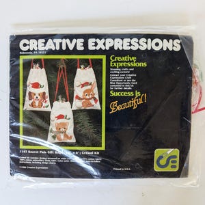 May include: A Creative Expressions 1147 Secret Pals Gift Bags 4 inch by 6 inch crewel kit in a clear plastic package. The black packaging features images of three white drawstring bags with embroidered animals wearing Santa hats and scarves.