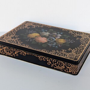 May include: A rectangular black metal tin featuring a central floral arrangement of pink, yellow, and blue flowers surrounded by a decorative gold filigree border that extends along the sides of the container lid and the base of the box.