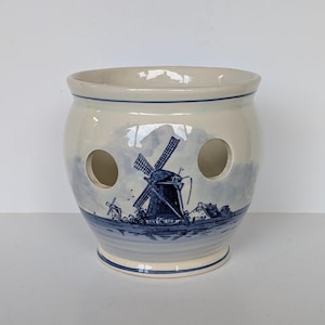 May include: Cream-colored ceramic planter with a blue windmill scene. The planter has two large circular holes and a blue rim. The windmill scene depicts a Dutch landscape with windmills and buildings. A decorative piece for home decor.